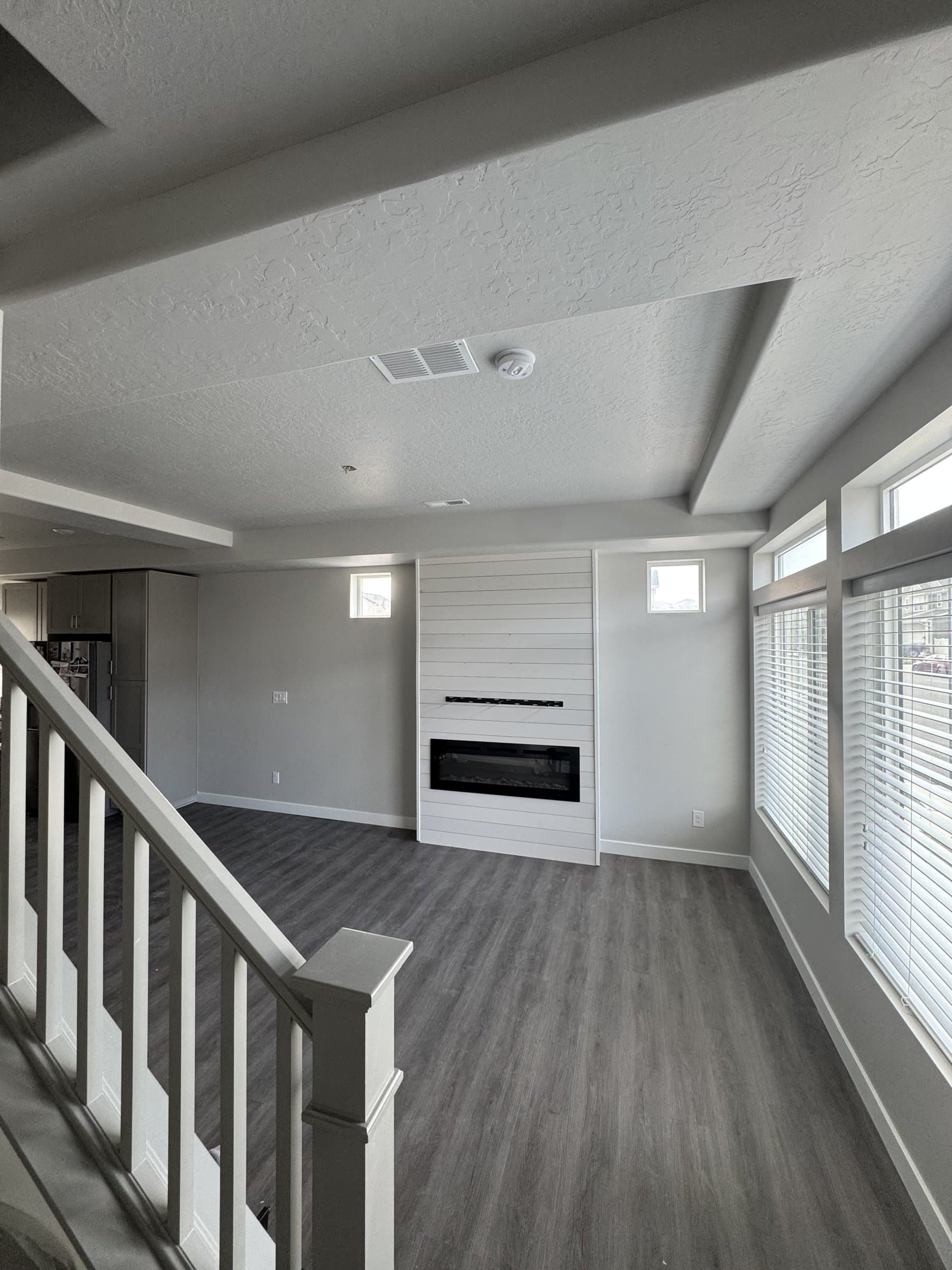 The finished basement living area from the top of the stairs: a white shiplap chimney breast with a linear electric fireplace, gray walls, gray plank flooring and the white painted stair rail in the foreground.