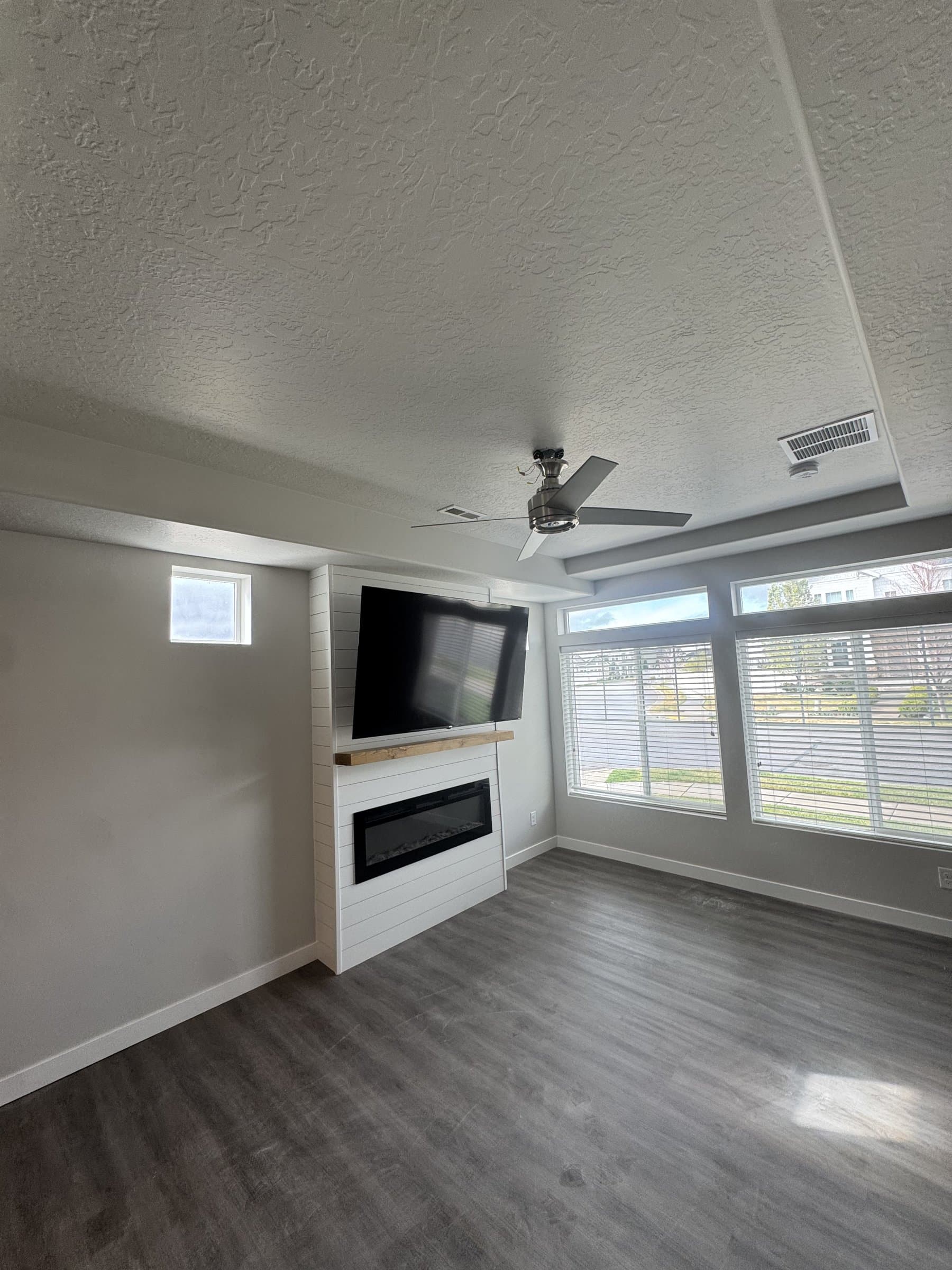 The finished basement living room with a television mounted over the shiplap fireplace on a wood mantel shelf, flat gray walls, gray plank flooring and daylight coming in from the window wall on the right.