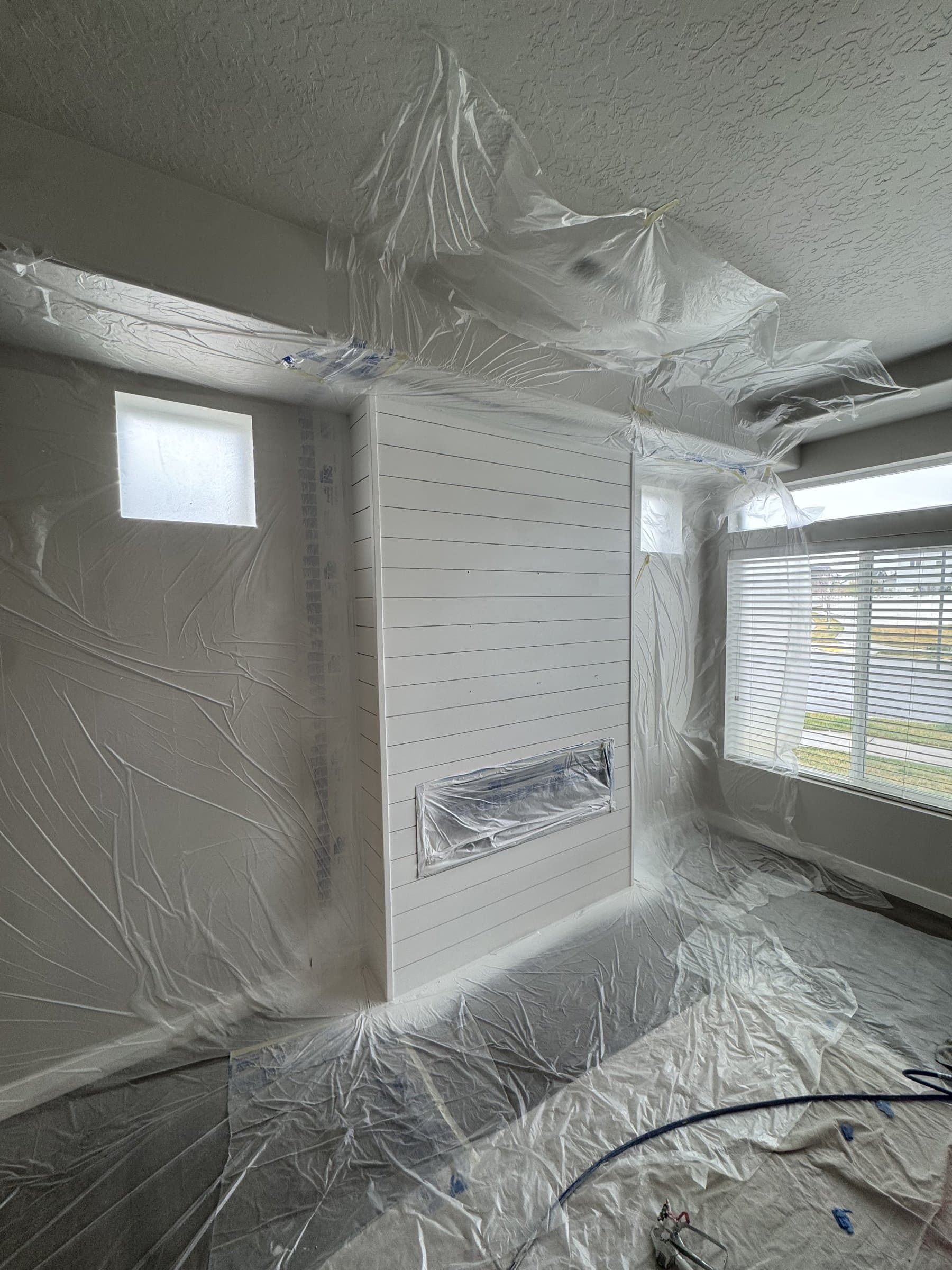 The basement feature wall part-way through, with plastic sheeting taped over the floor, the ceiling line and the fireplace opening, and the shiplap boards coated white ready for their finish coats.