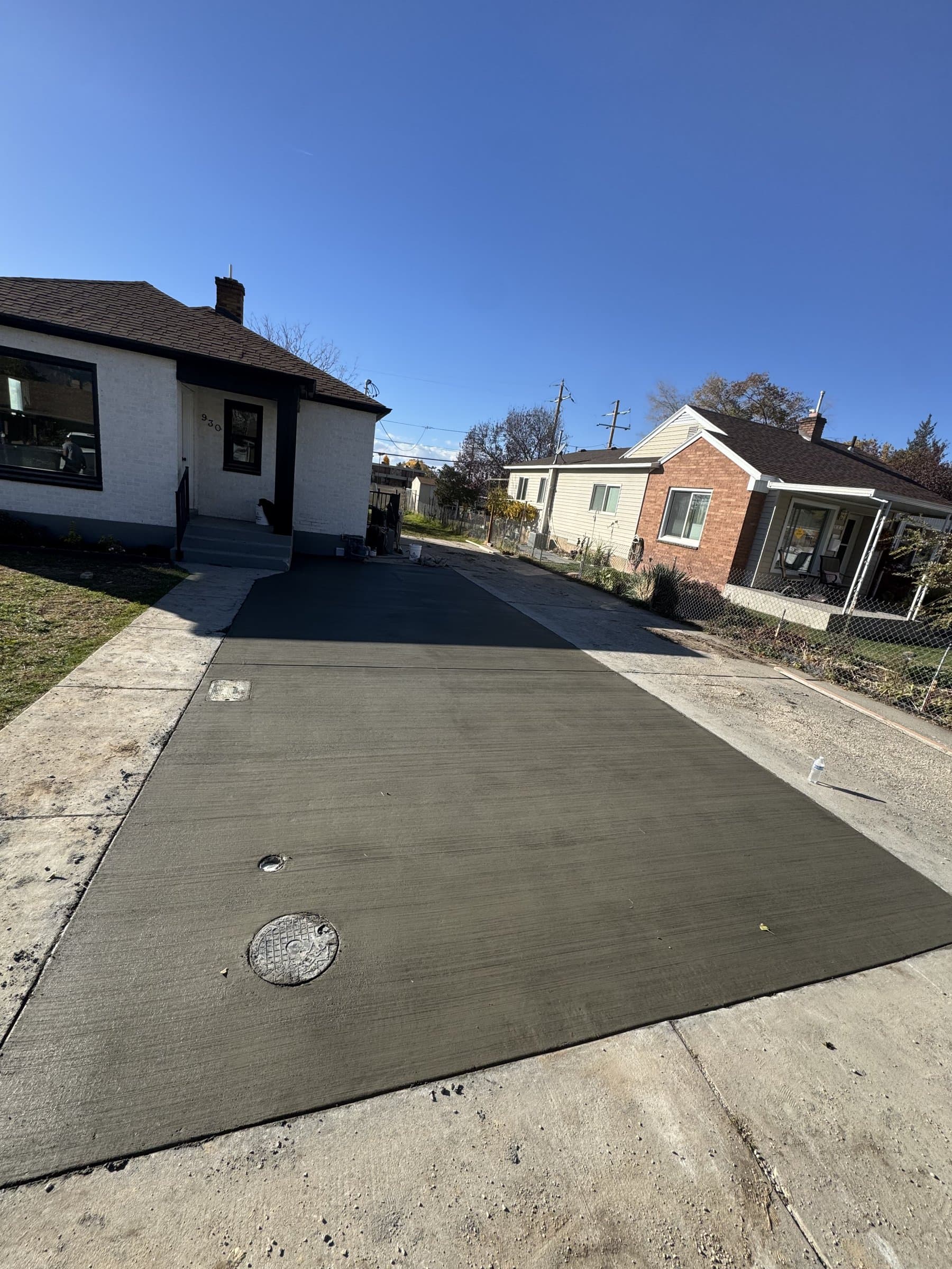 A newly poured concrete driveway running the full length of the house to the garage at the back, broom finished across its width with control joints cut in and a utility cover set flush into the slab.