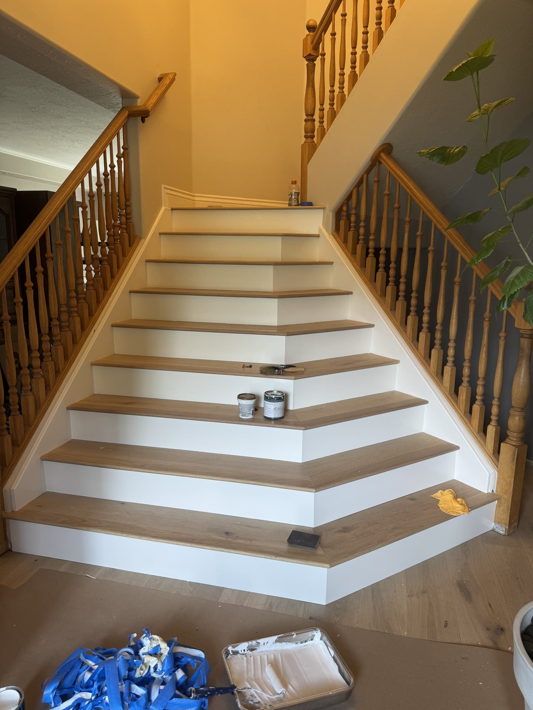 A staircase refinished with white painted risers and skirt against new oak treads, winding up past the original oak spindles and handrail, with a paint tray and used masking tape still on the floor at the bottom.